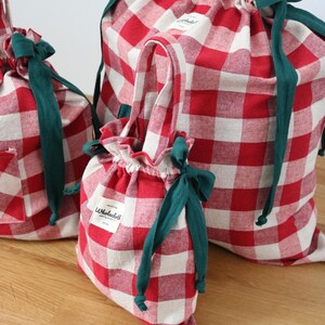 May include: Three red and white gingham drawstring bags of different sizes. Each bag features a green drawstring and a green ribbon bow. The smallest bag has a handle. The bags are on a wooden surface.