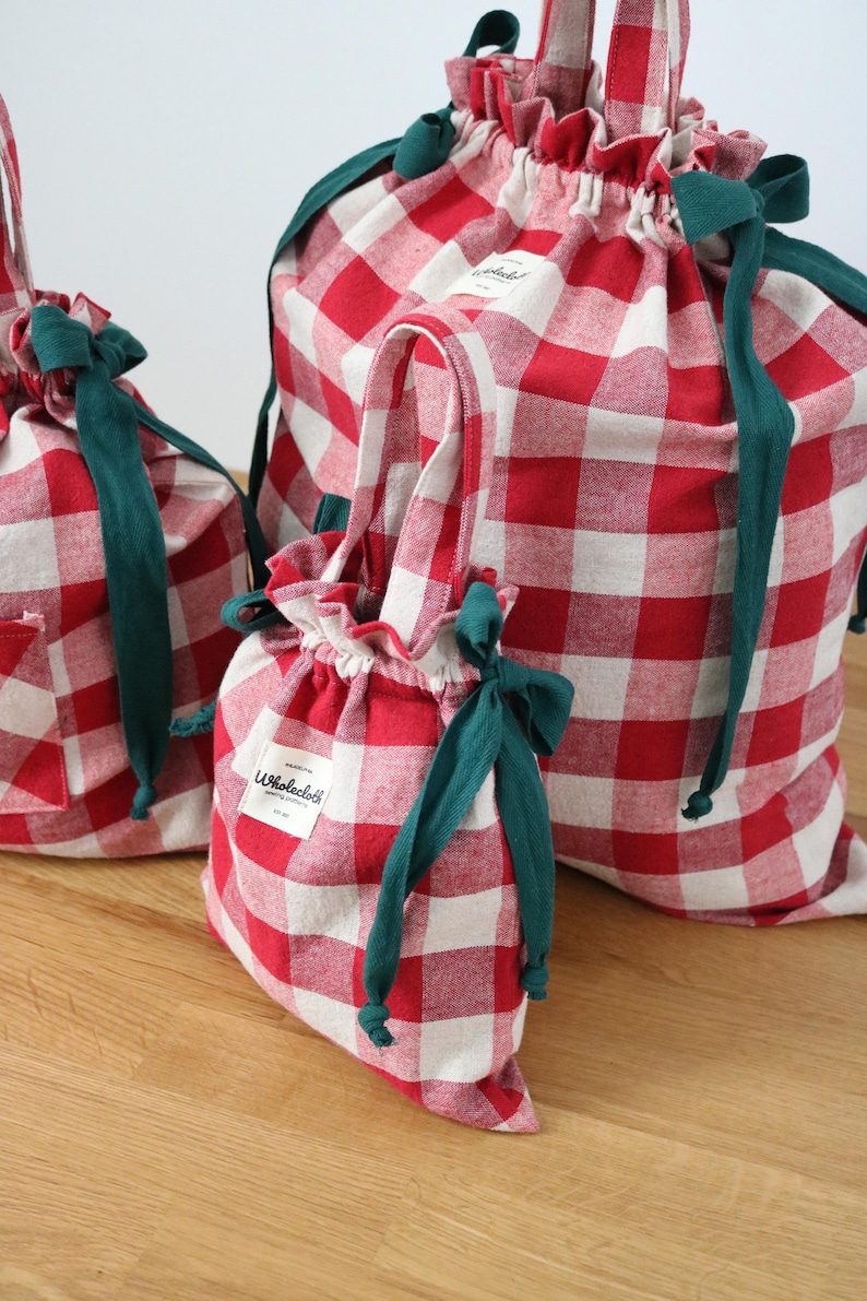 May include: Three red and white gingham drawstring bags of different sizes. Each bag features a green drawstring and a green ribbon bow. The smallest bag has a handle. The bags are on a wooden surface.