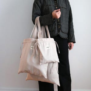 May include: Three beige linen tote bags of different sizes, with drawstring closures and small white labels. The bags are held by a person wearing a dark grey jacket and black trousers. The bags are designed for carrying items.