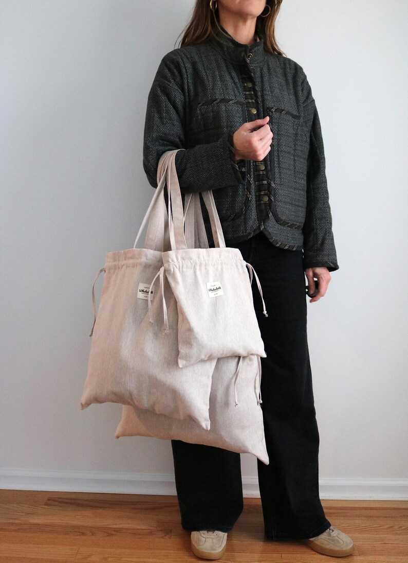May include: Three beige linen tote bags of different sizes, with drawstring closures and small white labels. The bags are held by a person wearing a dark grey jacket and black trousers. The bags are designed for carrying items.