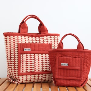 May include: Two red tote bags with matching handles and a front pocket. The larger bag features a cream-colored, semi-circle pattern. The smaller bag is solid red. Both bags have a small label that reads "Whalebell".