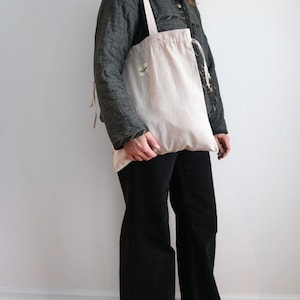 May include: A pale pink tote bag with a drawstring closure and a small label is held. The bag has a long strap and is made of a soft, natural fabric. The person is wearing a dark grey jacket and black trousers.