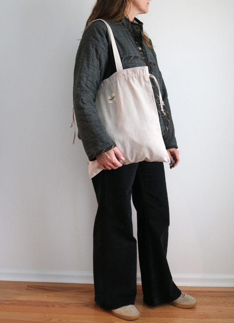 May include: A pale pink tote bag with a drawstring closure and a small label is held. The bag has a long strap and is made of a soft, natural fabric. The person is wearing a dark grey jacket and black trousers.