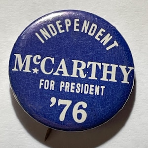 May include: A round, blue button with white text. The button reads "INDEPENDENT MCCARTHY FOR PRESIDENT '76". The button has a pin on the back and is likely a political campaign button.