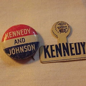 May include: Two vintage political campaign buttons. The larger button is white with blue lettering that reads "KENNEDY". The smaller button is red, white, and blue with the text "KENNEDY AND JOHNSON".