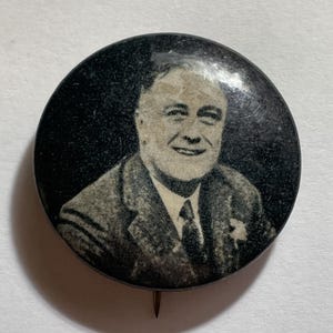 May include: A vintage black and white button featuring a portrait of a smiling man in a suit and tie. The button is round with a pin on the back. The image is a close-up of the man's face and upper body.