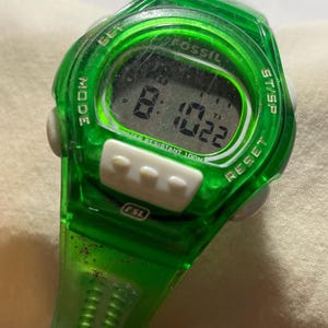 May include: A green Fossil digital watch with a rectangular display showing the time 8:10:22. The watch has white buttons and the text "WATER RESISTANT 100M". The watch is a sporty style.