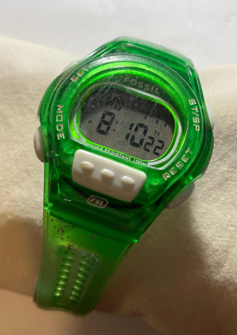 May include: A green Fossil digital watch with a rectangular display showing the time 8:10:22. The watch has white buttons and the text "WATER RESISTANT 100M". The watch is a sporty style.