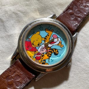 May include: A silver-toned wristwatch with a brown leather band. The watch face features a light blue background with illustrations of Winnie the Pooh, Tigger, and Piglet. The watch band is approximately 0.75 inches wide.