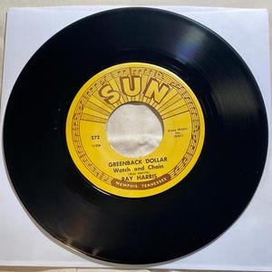 Puede incluir: Un disco de vinilo negro con una etiqueta amarilla. La etiqueta muestra la palabra "SUN" en una fuente estilizada, junto con el título de la canción "GREENBACK DOLLAR Watch and Chain" de Ray Harris, y la ubicación "MEMPHIS, TENNESSEE".