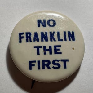 May include: A round, off-white button with the text "NO FRANKLIN THE FIRST" in dark blue block letters. The button has a pin on the back and shows signs of wear.