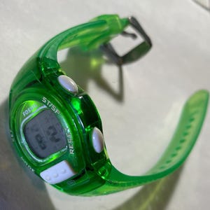 May include: A transparent green Fossil digital watch with a black digital display. The watch has white buttons and a green strap. The watch face displays the text "Fossil", "ST/SP", and "RESIST".