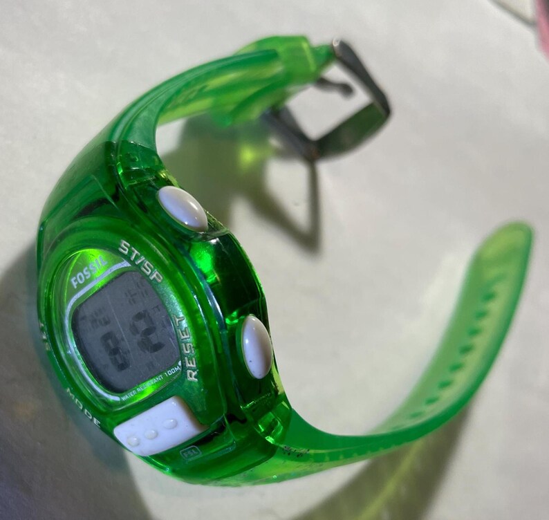 May include: A transparent green Fossil digital watch with a black digital display. The watch has white buttons and a green strap. The watch face displays the text "Fossil", "ST/SP", and "RESIST".