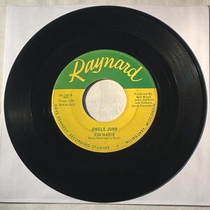 May include: A black vinyl record with a green and yellow label. The label reads "Raynard" in a stylized font, with the song title "Jingle Jump" by Kim Marie. The record is from Dave Kennedy Recording Studios.