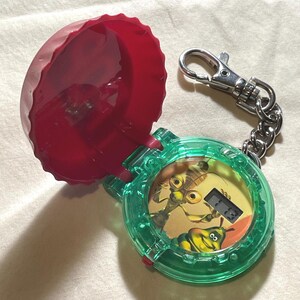 May include: A clear green toy watch with a red lid, featuring a cartoon insect design on the face. The watch has a digital display and a silver keychain attachment. The lid is open, revealing the watch face.