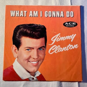 May include: Vintage vinyl record album cover featuring Jimmy Clanton. The album cover is orange with the text "WHAT AM I GONNA DO" at the top. The singer's name, "Jimmy Clanton", is written in cursive. The image shows a smiling man with dark hair and blue eyes.