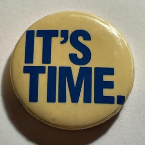 May include: A round, cream-colored button with the text "IT'S TIME." in bold, blue capital letters. The button has a glossy finish and a slightly worn appearance. The text is centered on the button.