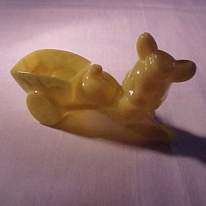 May include: A yellow glass figurine of a horse pulling a wagon. The horse has a long neck and a long tail. The wagon has a rounded shape and two wheels.