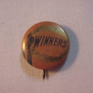 May include: A round, brown button with a gold-colored design and the word "WINNERS" printed in black letters. The button has a pin on the back.