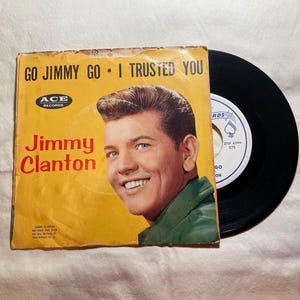 May include: Vintage 45 RPM record featuring Jimmy Clanton. The record sleeve is yellow with the text "GO JIMMY GO • I TRUSTED YOU" and a portrait of Jimmy Clanton. The record is black.