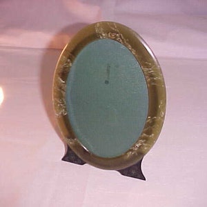 May include: An oval-shaped picture frame with a green and white marbled finish. The frame has a dark green mat and a black stand.