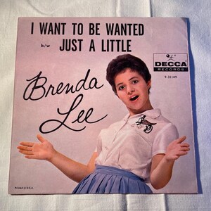 May include: Vintage vinyl record cover featuring Brenda Lee. The cover has the text "I WANT TO BE WANTED" and "JUST A LITTLE". Brenda Lee is pictured wearing a white shirt and blue skirt. The Decca Records logo is in the upper right corner.