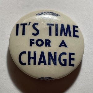 May include: A round, off-white button with the text "IT'S TIME FOR A CHANGE" in dark blue. The button has a glossy finish and appears to be vintage. The text is centered on the button.