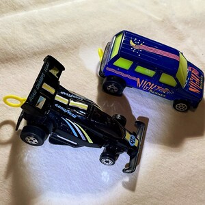 May include: A black Road Champs toy race car with Goodyear branding and a yellow pull-ring, alongside a blue Night Runner toy van featuring yellow windows, a moon and stars graphic on the roof, and a yellow pull-ring attached to the rear.