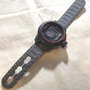 May include: A black and gray toy watch with a red accent ring around the face. The watch has a digital display and a gray band with a unique, textured design. The watch band measures approximately 9 inches in length.