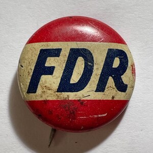 May include: A vintage round button with the letters "FDR" in navy blue on a cream-colored band, set against a red background. The button has a pin on the back and shows signs of age.