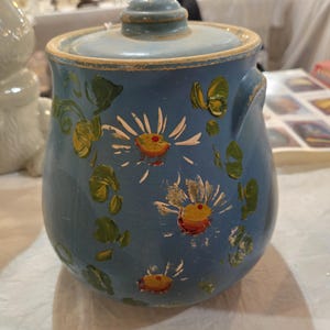 May include: A blue ceramic jar with a lid, adorned with hand-painted white flowers with yellow centers and green leaves. The jar has two small handles and a matching blue lid. A vintage kitchenware item.