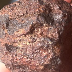 Pode incluir: Close-up de uma rocha áspera e texturizada com uma cor marrom avermelhada. A superfície é irregular, com pequenas protuberâncias irregulares e uma aparência desgastada. A cor da rocha varia do marrom escuro a tons enferrujados mais claros.