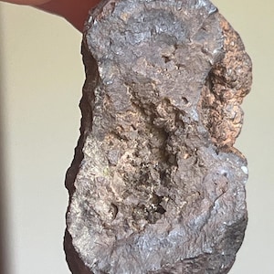 Könnte beinhalten: Ein dunkelbrauner, unregelmäßig geformter Stein mit einer rauen, strukturierten Oberfläche. Der Stein hat eine Mischung aus dunklen und helleren Brauntönen, wobei einige Bereiche metallischer erscheinen. Die Gesamtform ist etwas länglich, mit ein paar kleinen Löchern.