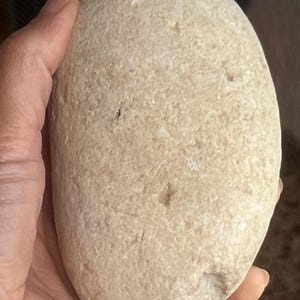 May include: A smooth, oval-shaped, light beige stone. The stone is held in a hand, showcasing its natural texture and colour variations. The stone's surface has small imperfections and subtle colour differences.
