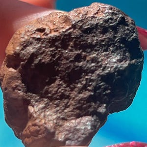 Puede incluir: Una pequeña roca de forma irregular con una superficie de color marrón oscuro, porosa y texturizada. El objeto presenta un exterior rugoso e irregular con hendiduras profundas y un acabado mate, mostrando una composición densa y granular.