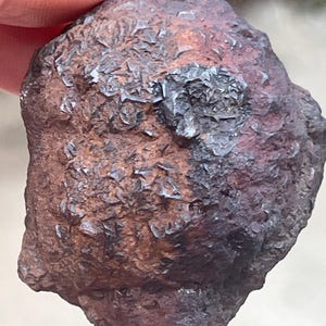 Può includere: Una roccia irregolare di colore marrone rossiccio scuro con una superficie ruvida. Presenta formazioni cristalline su un lato, con una lucentezza metallica. L'aspetto generale suggerisce un campione minerale naturale.