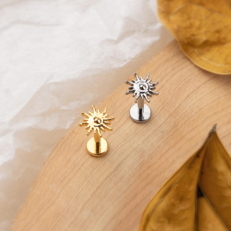May include: Two sun-shaped lip piercing studs. One is gold-colored, the other is silver-colored. Each stud features a sunburst design and a flat back. They are displayed on a wooden surface with dried leaves.