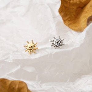 May include: Two sun-shaped stud earrings. One earring is gold-colored, and the other is silver-colored. The earrings are small, with a detailed sunburst design. They are placed on a white, textured surface.