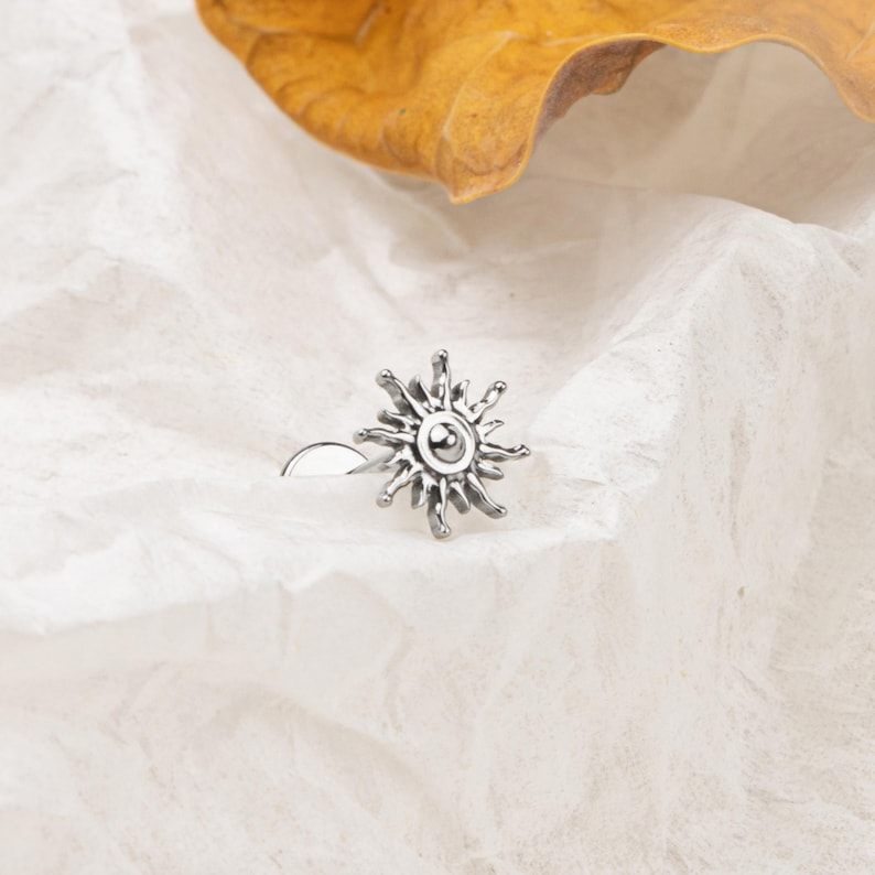 May include: A silver-colored nose ring featuring a sun design. The sun has a detailed, textured surface with rays extending outwards from a central circle. The jewelry is set against a white, textured background with a dried leaf in the upper corner.