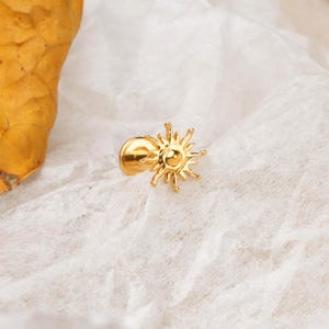 May include: A gold-colored sun-shaped pin. The pin features a circular center with radiating lines, resembling sun rays. The pin is set against a white, textured background, with a portion of a yellow leaf visible.