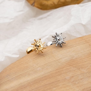 May include: Two sun-shaped cufflinks, one gold-toned and one silver-toned, rest on a wooden surface. The cufflinks feature a detailed sunburst design with a central circular element. The background includes a white, textured fabric.