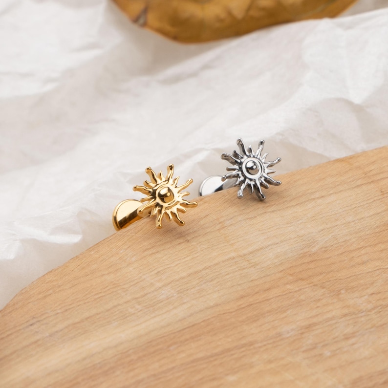 May include: Two sun-shaped cufflinks, one gold-toned and one silver-toned, rest on a wooden surface. The cufflinks feature a detailed sunburst design with a central circular element. The background includes a white, textured fabric.