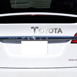 May include: A white Tesla Model X with a silver Toyota emblem on the back. The emblem is placed above the license plate.