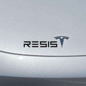 Op de afbeelding: Een witte auto met een zwart Tesla-logo en een zwarte vinyl sticker met de tekst "RESIS" op de motorkap.