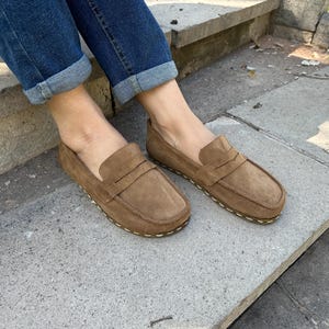 Puede incluir: Mocasines de ante marr&oacute;n con un dise&ntilde;o cl&aacute;sico de mocas&iacute;n. Los zapatos tienen una puntera redondeada y una suela cosida. La imagen muestra los zapatos sobre una superficie de hormig&oacute;n, con vaqueros azules visibles.