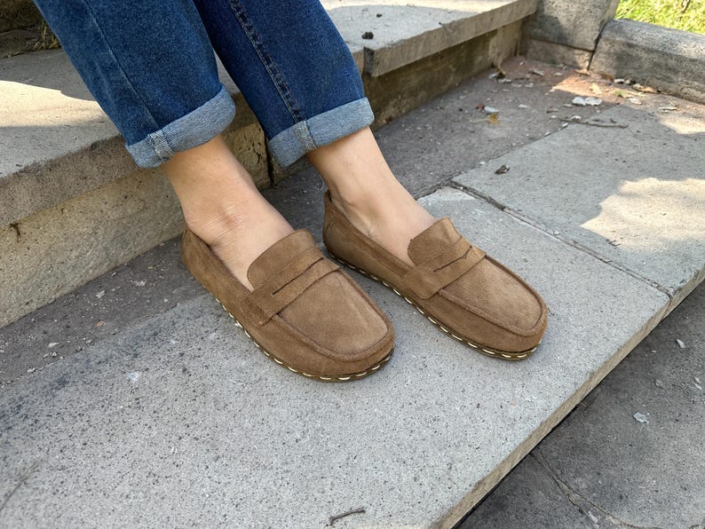 Puede incluir: Mocasines de ante marr&oacute;n con un dise&ntilde;o cl&aacute;sico de mocas&iacute;n. Los zapatos tienen una puntera redondeada y una suela cosida. La imagen muestra los zapatos sobre una superficie de hormig&oacute;n, con vaqueros azules visibles.