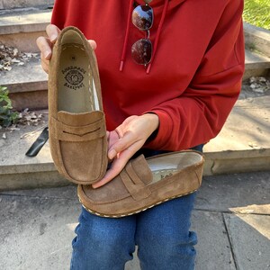 Puede incluir: Mocasines de ante marr&oacute;n con un dise&ntilde;o cl&aacute;sico de penny loafer. Los zapatos tienen una suela cosida y un peque&ntilde;o logotipo en relieve en el interior. Los zapatos son de un color c&aacute;lido y neutro.
