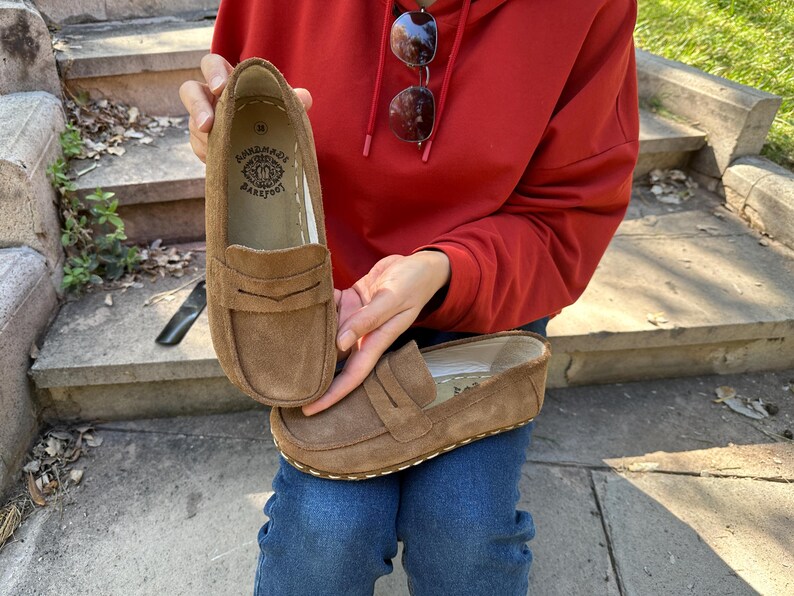 Puede incluir: Mocasines de ante marr&oacute;n con un dise&ntilde;o cl&aacute;sico de penny loafer. Los zapatos tienen una suela cosida y un peque&ntilde;o logotipo en relieve en el interior. Los zapatos son de un color c&aacute;lido y neutro.