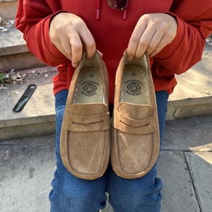 Puede incluir: Un par de mocasines de ante marr&oacute;n con un dise&ntilde;o cl&aacute;sico de mocas&iacute;n con borlas. Los zapatos son sostenidos por una persona que lleva una sudadera con capucha roja y vaqueros azules. Las plantillas est&aacute;n estampadas con un logotipo "HANDMADE BAREFOOT".