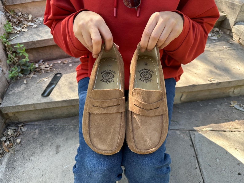 Puede incluir: Un par de mocasines de ante marr&oacute;n con un dise&ntilde;o cl&aacute;sico de mocas&iacute;n con borlas. Los zapatos son sostenidos por una persona que lleva una sudadera con capucha roja y vaqueros azules. Las plantillas est&aacute;n estampadas con un logotipo "HANDMADE BAREFOOT".